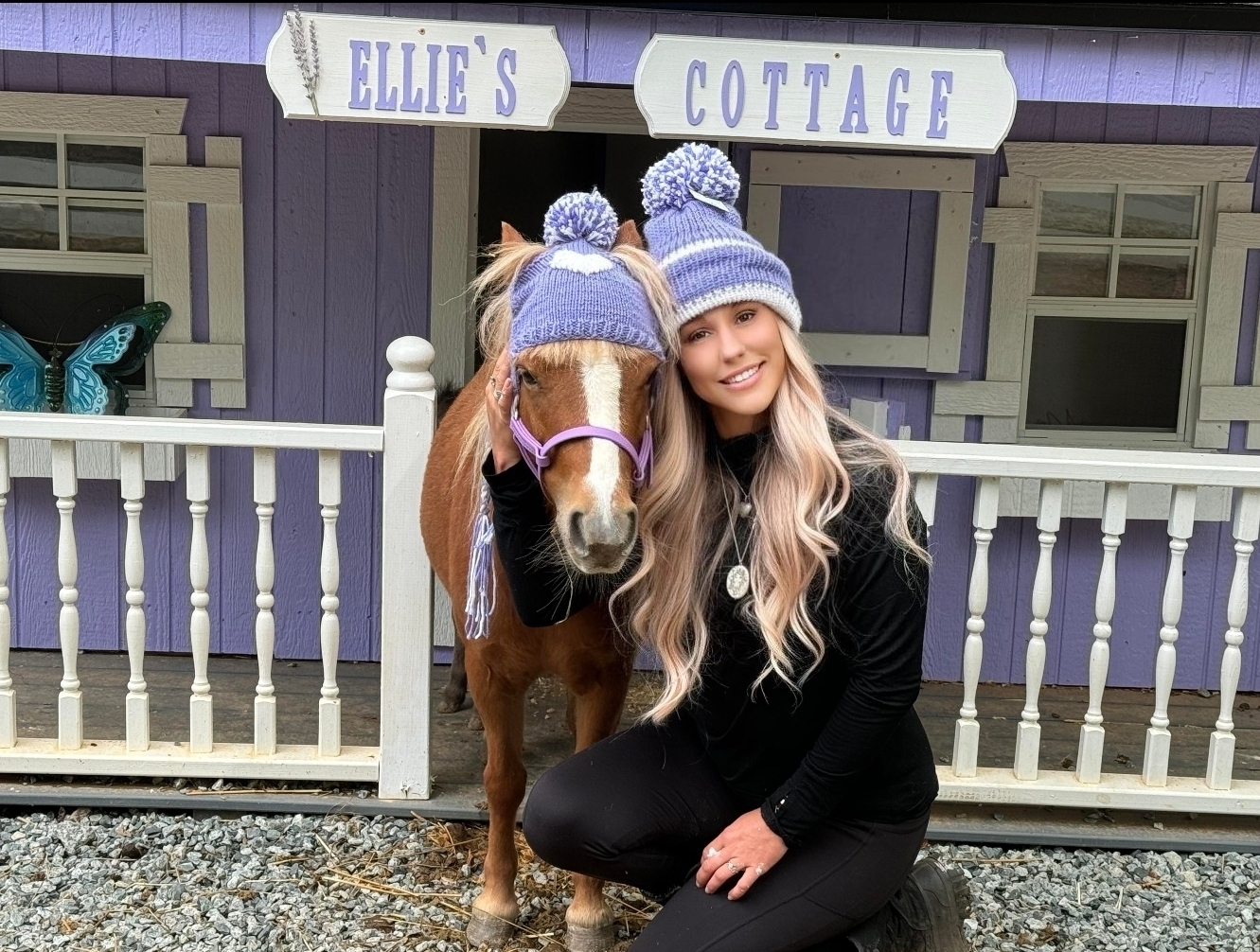 Danielle with Ellie the miniature horse in matching purple knit hats in front of Ellie's Cottage